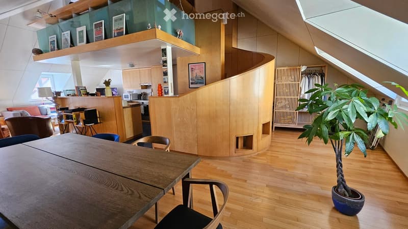 Open-plan living and dining area with wooden flooring, a large dining table with chairs, a sofa, a kitchen area with cabinets and appliances, and a potted plant near the window.
