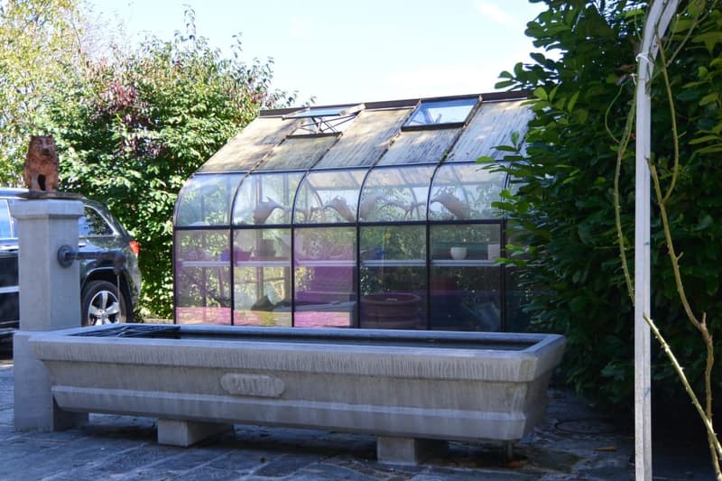 A garden area features a stone fountain in the foreground and a greenhouse with glass panels and a slanted roof in the background, surrounded by lush greenery and trees.