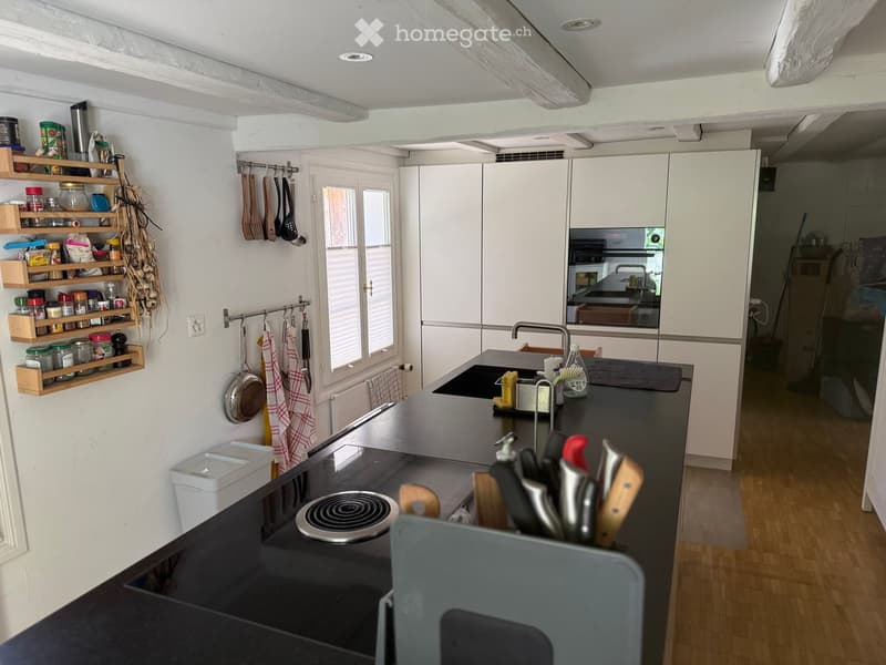Modern kitchen with white cabinets, black countertops, induction cooktop, sink, and various kitchen utensils. Wooden shelves hold jars and spices. A window provides natural light.