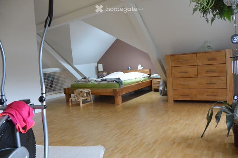 Cozy attic bedroom with wooden floors, slanted ceilings, a double bed with green bedding, a wooden chest of drawers, a small stool, and a hanging plant.