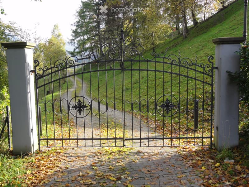 Ornate black metal gate with decorative scrollwork and circular motifs, flanked by grey stone pillars, leading to a gravel pathway surrounded by autumn leaves and green grass.