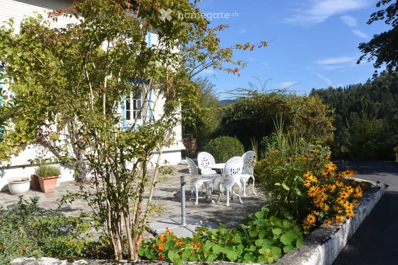 A well-maintained garden area with a white table and chairs, surrounded by colorful flowers and greenery, adjacent to a white house with blue trim.