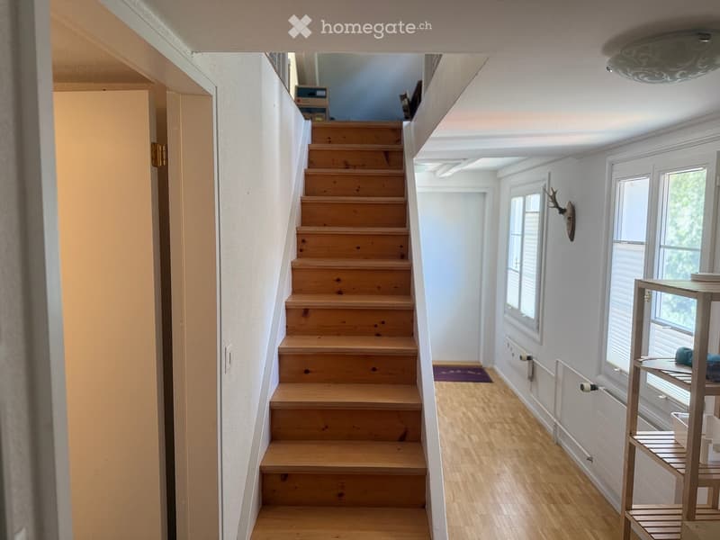 Wooden staircase leading to an open area with white walls, large windows, and a small shelf with items on the right side.