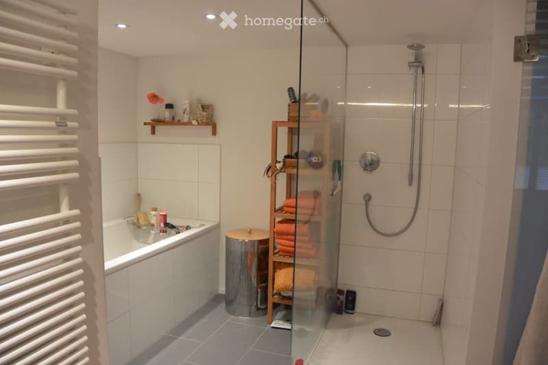 Modern bathroom with a bathtub, glass-enclosed shower, white tiled walls, and a wooden shelf holding towels and toiletries.