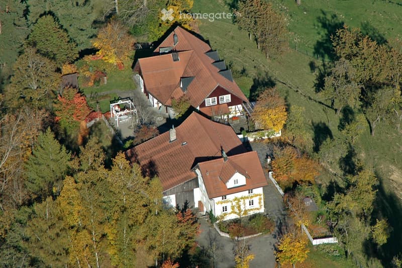 Aerial view of a two-story house with red-tiled roofs, surrounded by trees and greenery. The house features multiple chimneys and is situated in a rural, forested area.
