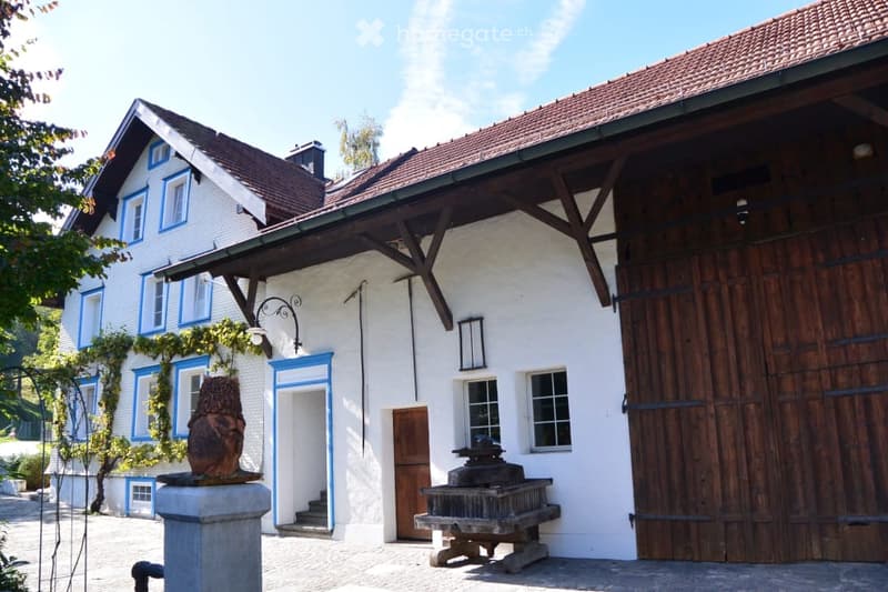 Traditional two-story building with white walls, blue window frames, wooden barn-style door, and a small fountain in front.