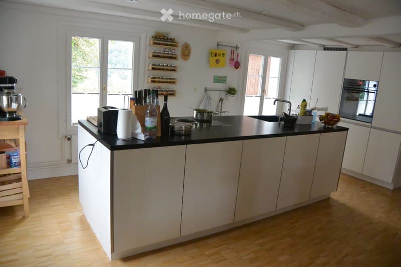 Modern kitchen with white cabinets, black countertop, and wooden flooring. Features include a kitchen island, sink, oven, and various appliances and utensils.