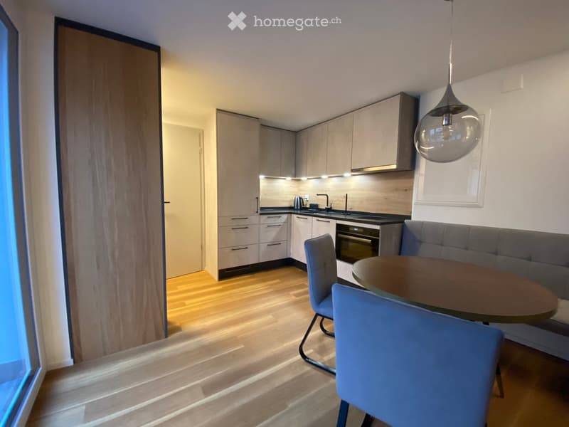 Modern kitchen and dining area with light wood flooring, grey sofa, round dining table with blue chairs, and sleek kitchen cabinets.