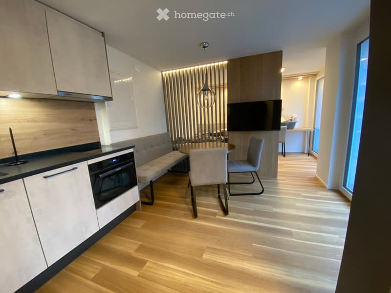 Modern, open-plan living and dining area with light wood flooring, white cabinets, a built-in oven, a bench seating, a dining table with chairs, and a television mounted on the wall.