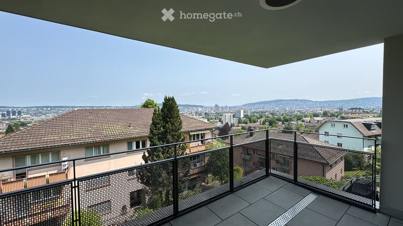 Balcony with a metal railing, overlooking a cityscape with houses, trees, and distant mountains.