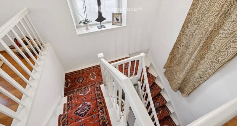 Spacious staircase with wooden steps, white railings, and patterned red runner. A window with a small plant and decorative items is visible above the stairs. The walls are painted white, and a patterned curtain hangs on the right side.