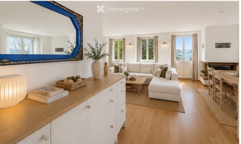 Bright living room with white walls, large windows, and a view. Features a white sectional sofa, coffee table, side table, mirror, vase with greenery, books, and a dining area with chairs and table.