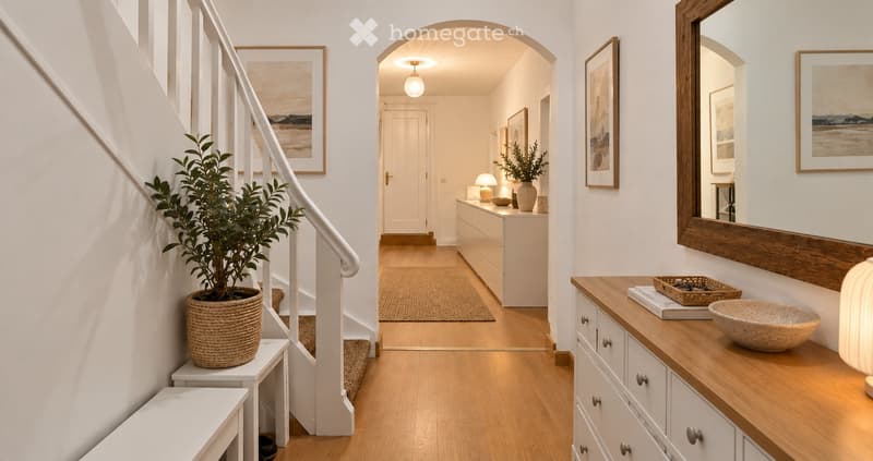 Bright hallway with wooden flooring, white walls, and a staircase leading upwards. Features include a potted plant on a small table, a large mirror, a console table with a bowl and books, and framed artwork on the walls.