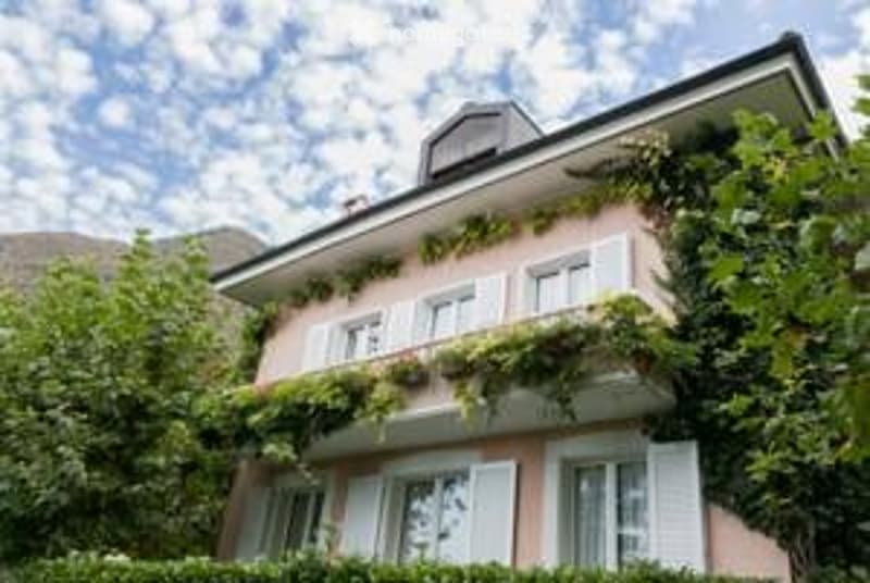 A charming two-story house with white-framed windows, light pink walls, and a lush green ivy-covered facade. The roof is dark, and the house is surrounded by trees and greenery.