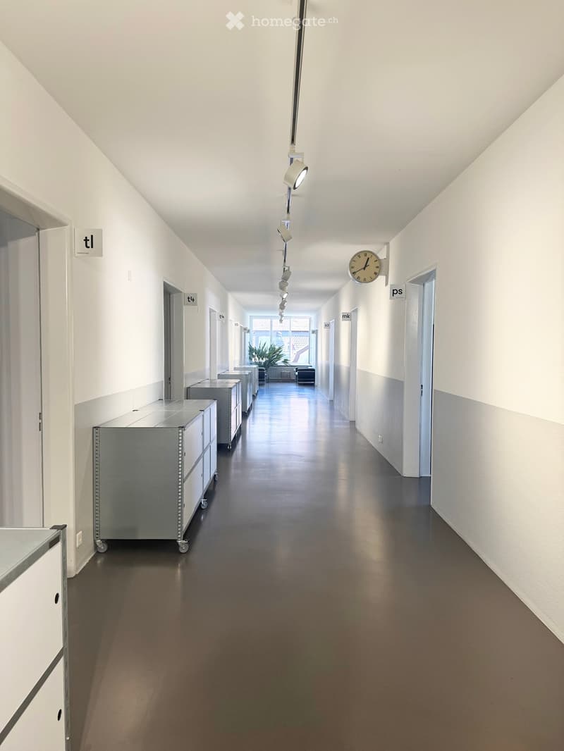 Clean, modern hallway with white walls, dark flooring, and a series of metal cabinets on the left side. A clock is mounted on the right wall, and several doors are visible along the corridor.