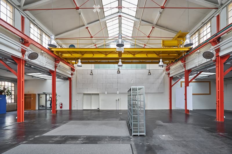 Large industrial-style space with high ceilings, exposed red steel beams, large windows, and industrial lighting. The floor is concrete, and there are minimal furnishings, including a metal shelving unit and a small cabinet.