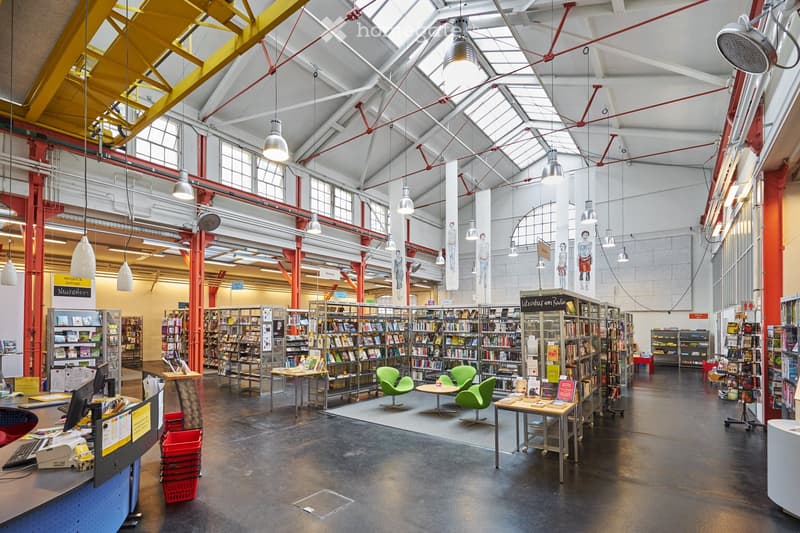 A spacious, modern library with high ceilings, exposed steel beams, and large windows. The area features tables, chairs, bookshelves filled with books, and a reception desk with a computer and printer.