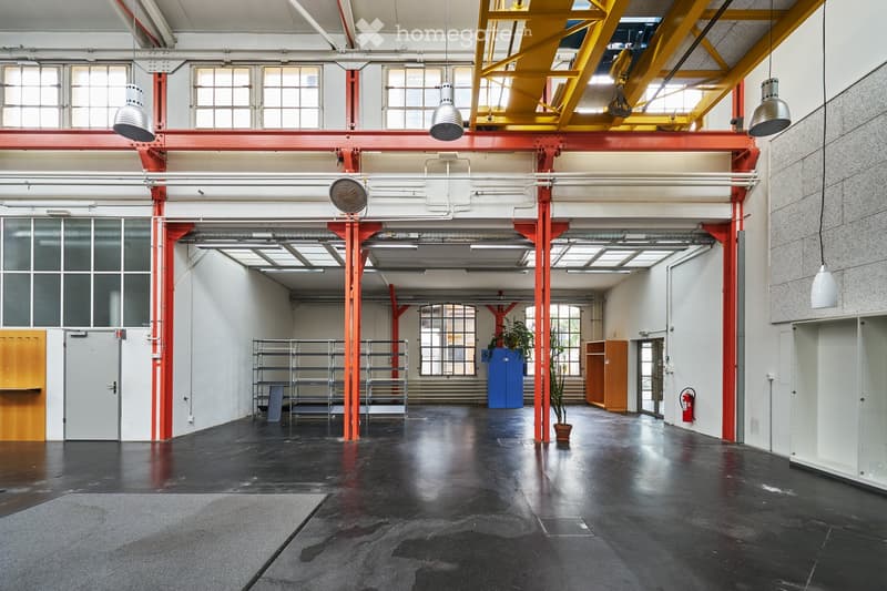 Large, open industrial-style space with high ceilings, exposed red steel beams, large windows, and a polished black concrete floor. Minimal furnishings include metal shelving units, a potted plant, and a fire extinguisher.