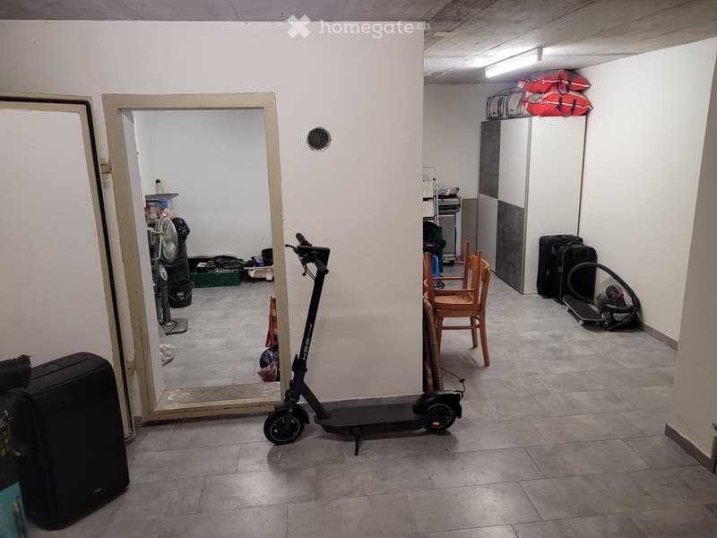A small, furnished room with tiled flooring and white walls. Contains a scooter, a wooden chair, a suitcase, a fan, a skateboard, and a large mirror.