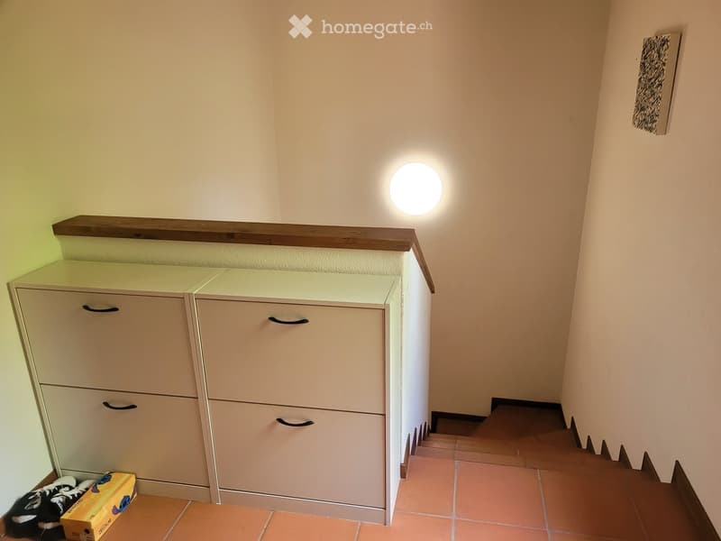 A narrow hallway with beige walls, a round wall-mounted light fixture, a built-in storage unit with drawers, and a small wooden shelf above it. The floor is tiled in warm orange tiles.