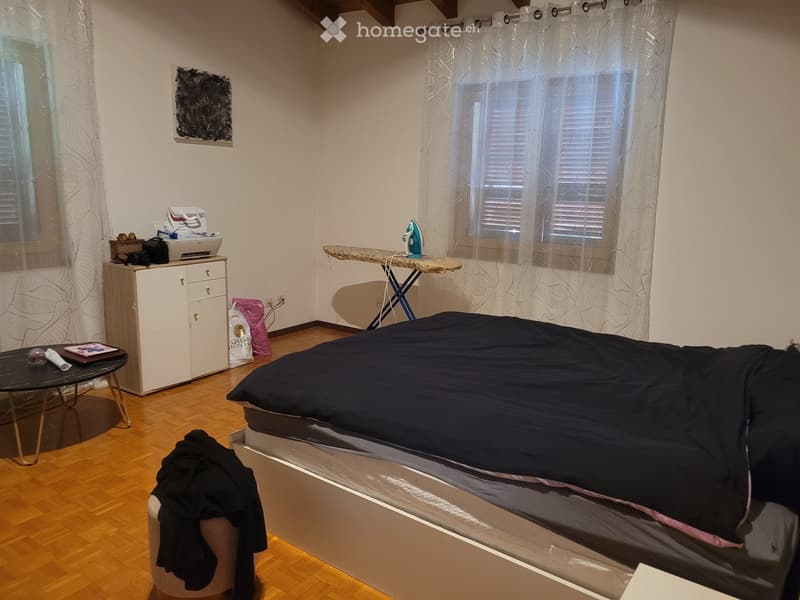 Bedroom with a bed, small round table, cabinet, ironing board, and mirror. Wooden flooring and white walls.