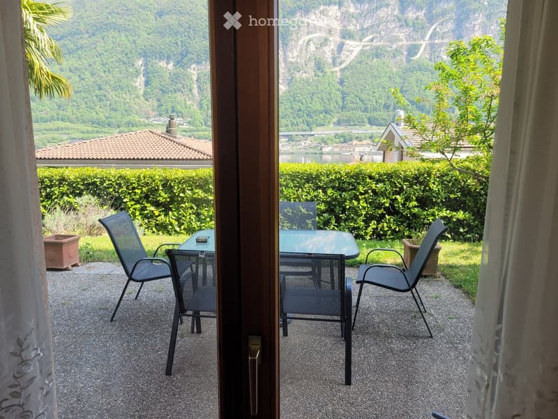 Balcony with a scenic mountain view, featuring a table and chairs, surrounded by greenery and a hedge.