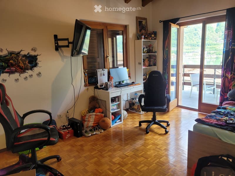 A furnished room with a wooden floor, a desk with a computer, a gaming chair, a bed with a colorful blanket, a shelf with various items, and a large window with a view of the outdoors.
