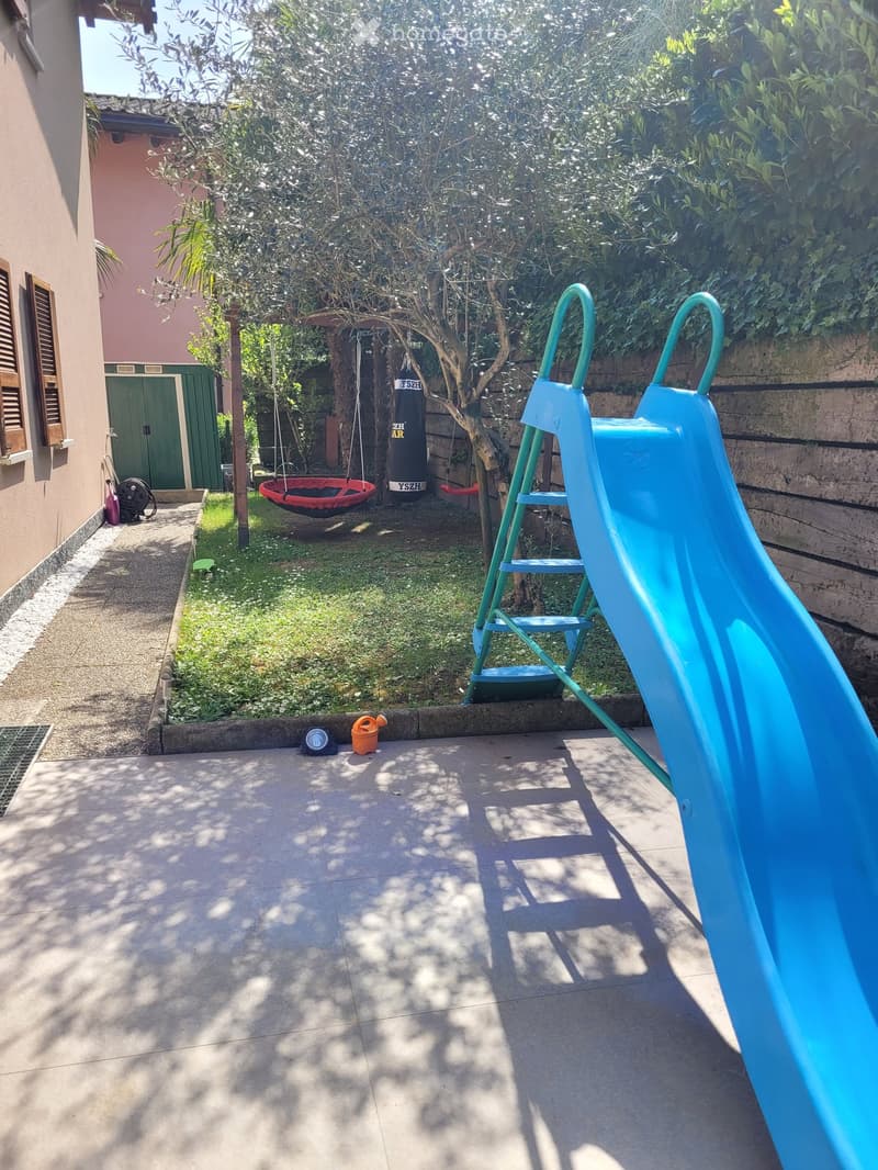 Garden area with a blue slide, swing, punching bag, and small tree. Concrete patio with shadows from surrounding foliage.