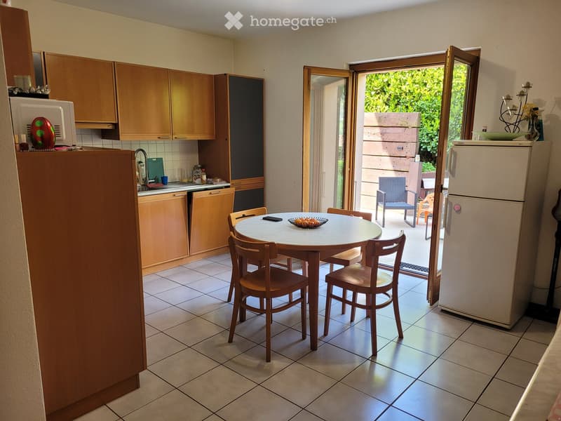 The kitchen features wooden cabinets, a white refrigerator, a microwave, a sink with a faucet, a dishwasher, and a round wooden dining table with four chairs. A sliding glass door opens to a patio with outdoor furniture.