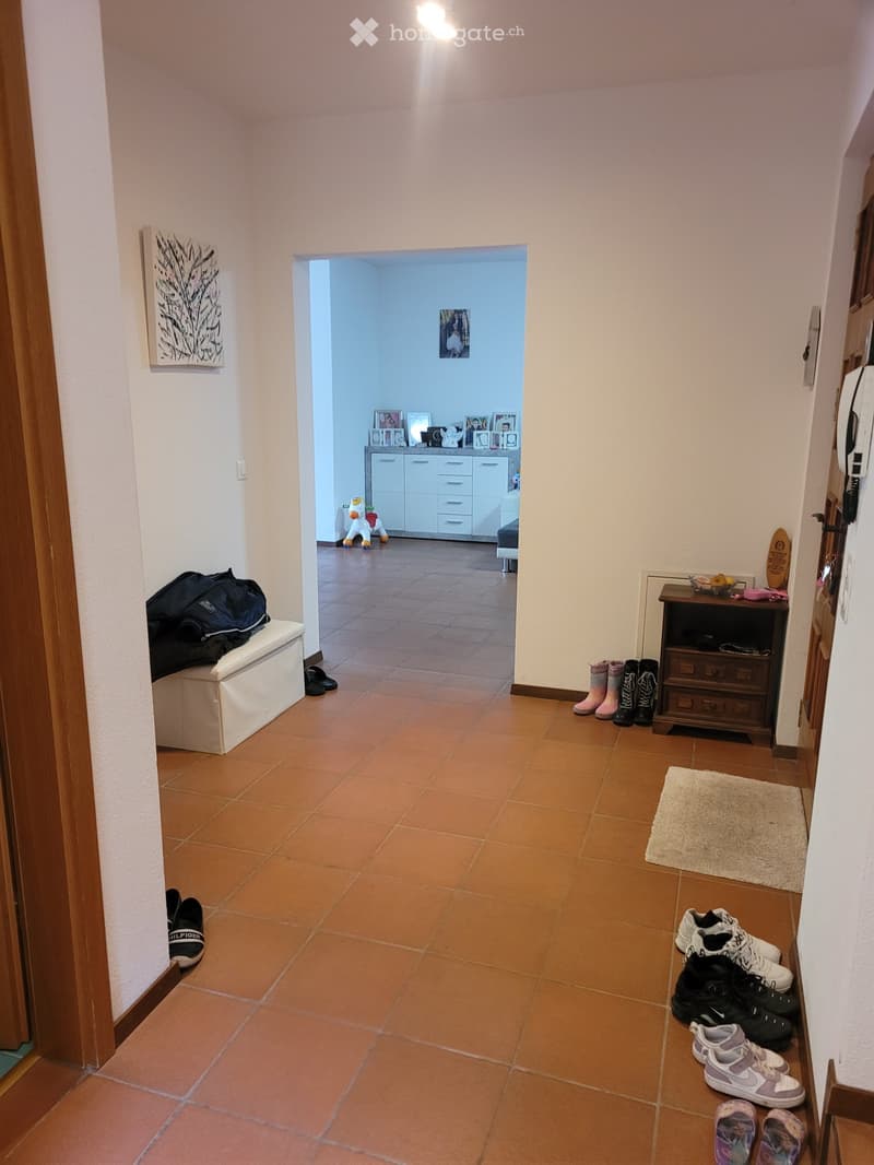 A well-lit hallway with terracotta tiled flooring, leading to an adjacent room. The hallway features a small bench with a backpack, several pairs of shoes and boots placed along the walls, and a small wooden cabinet with additional items. The adjacent roo