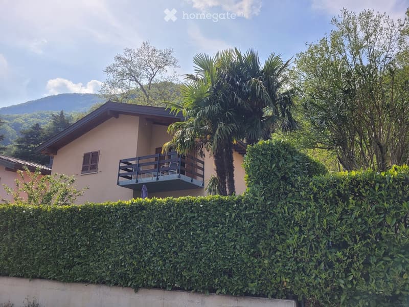 Two-story house with a balcony, surrounded by lush greenery and trees, with a well-maintained hedge in the foreground.