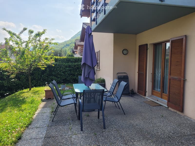 Outdoor patio area with a glass table, four chairs, and a folded umbrella, adjacent to a house with wooden shutters and a view of greenery.