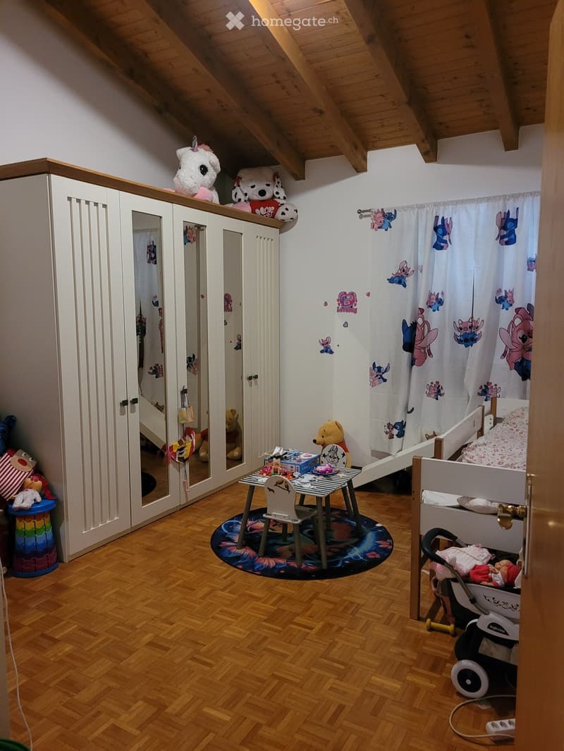 Cozy children's bedroom with wooden flooring, slanted ceiling, wardrobe, small table with chairs, toys, and a baby stroller.