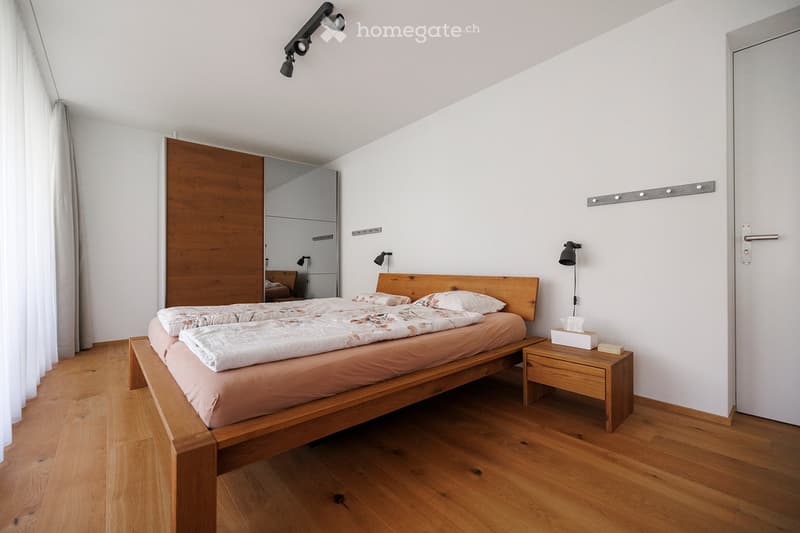 Modern bedroom with wooden bed, nightstand, and wardrobe. Features wooden flooring, white walls, and a large mirror on the wardrobe.