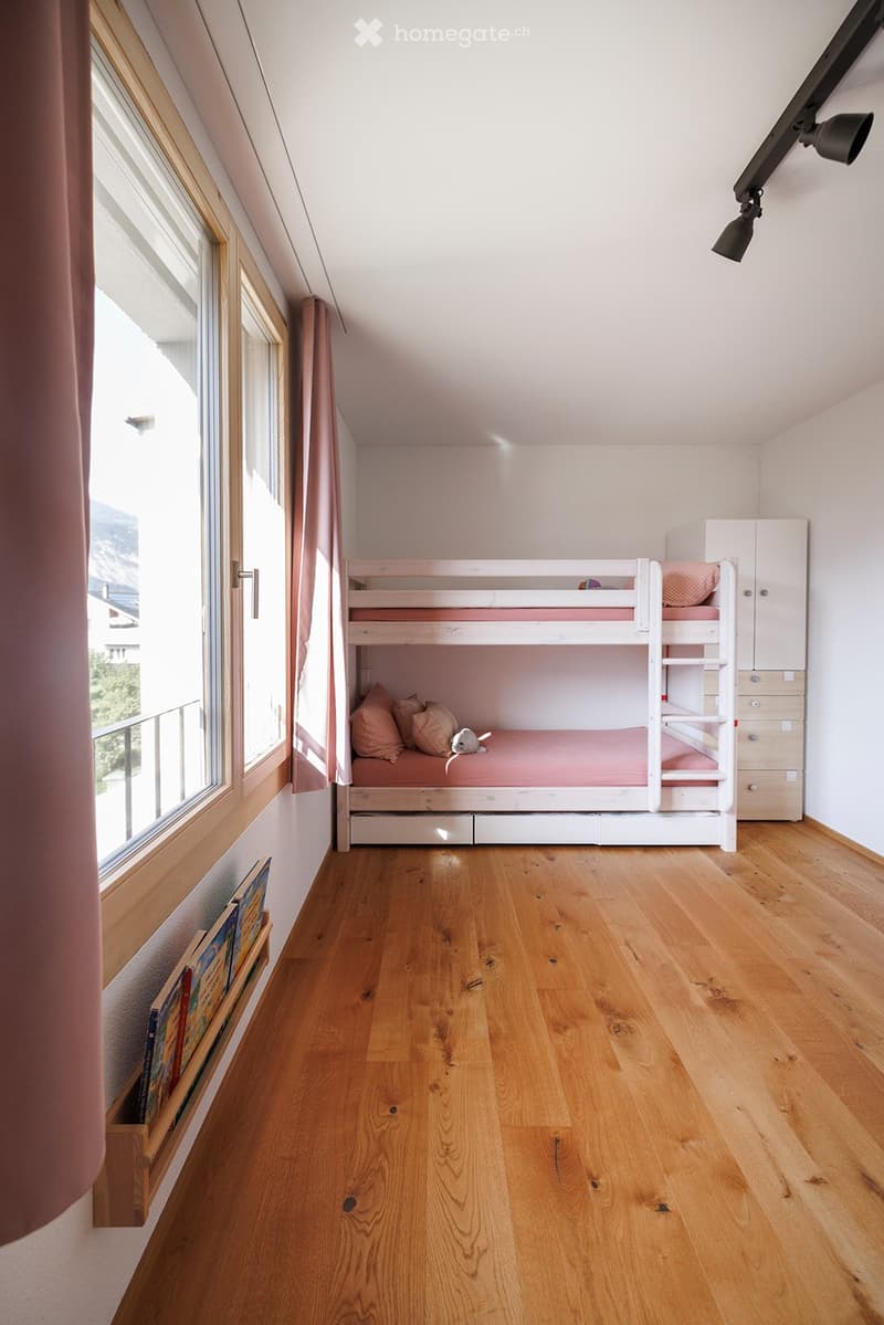 Cozy bedroom with wooden floor, bunk bed, pillows, and a small cabinet. Large windows with curtains provide natural light.