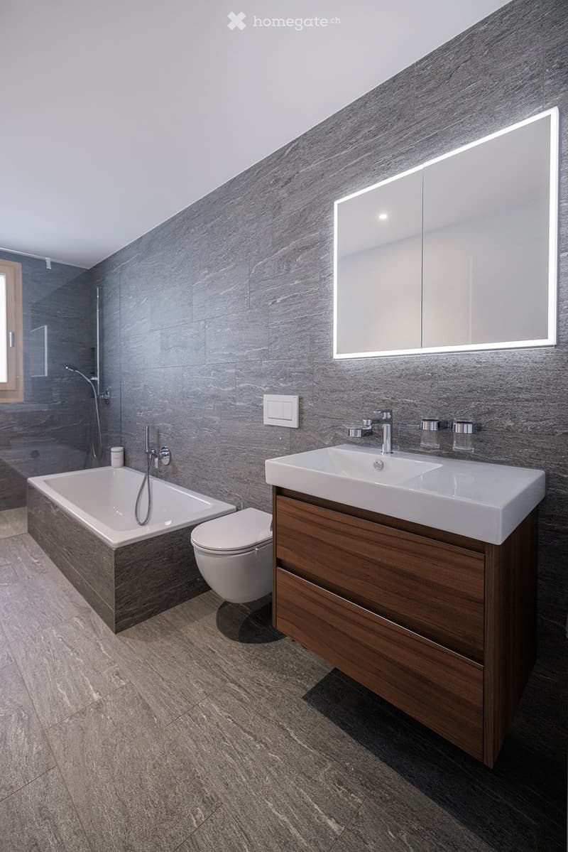 Modern bathroom with a dark grey tiled wall, a white bathtub, a toilet, a wooden vanity with a sink, and a large illuminated mirror above the sink.