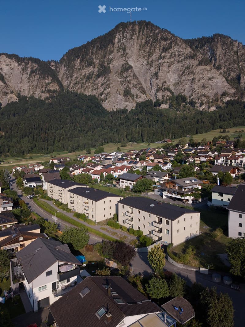 Aerial view of a residential area nestled in a valley surrounded by mountains, featuring multiple houses and apartment buildings with various roof styles and solar panels.