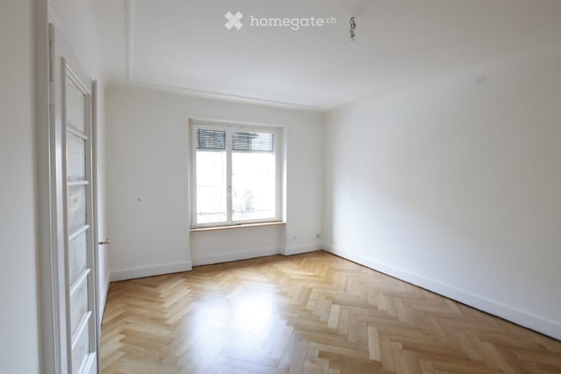 Completely empty room with white walls, wooden herringbone flooring, and a single window with blinds.