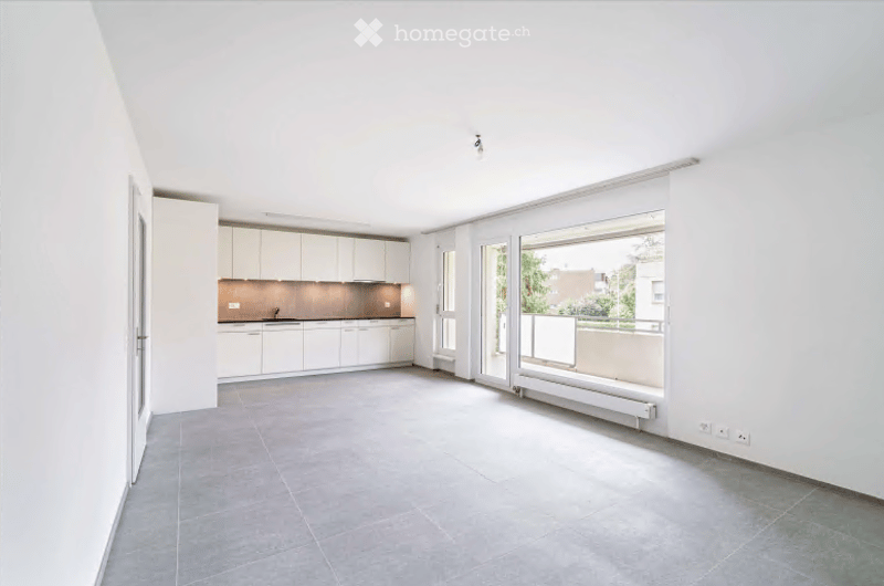 Open-plan space with a modern kitchen area featuring white cabinets and a sink, connected to a living area with large windows and a balcony access. The floor is tiled, and the walls are painted white.