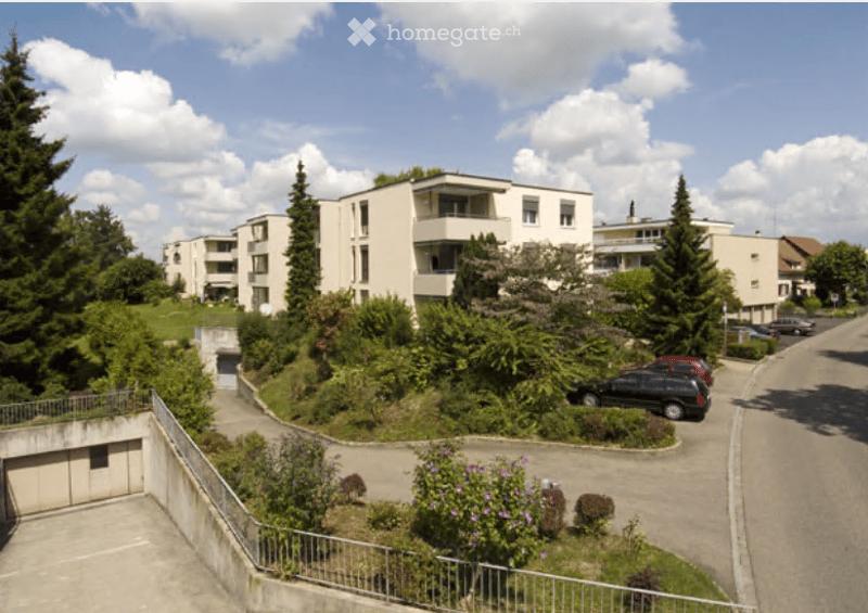 Multi-story residential building with balconies, surrounded by greenery and parking area.