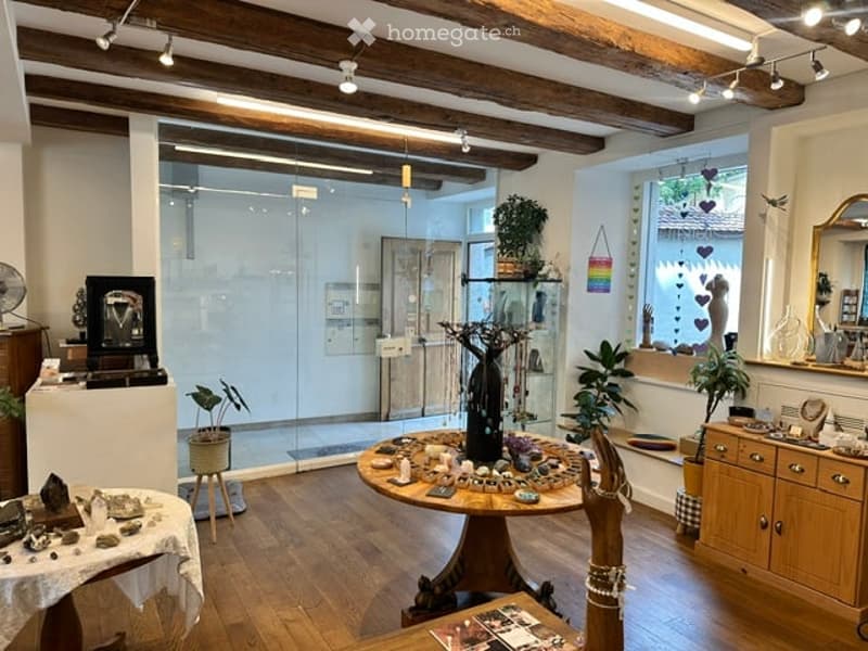 A cozy, well-lit room with wooden floors and exposed wooden beams. The space features a round wooden table with various decorative items, a small stool, a cabinet with drawers, a fan, a mirror, and several plants. The room also includes a glass display ca