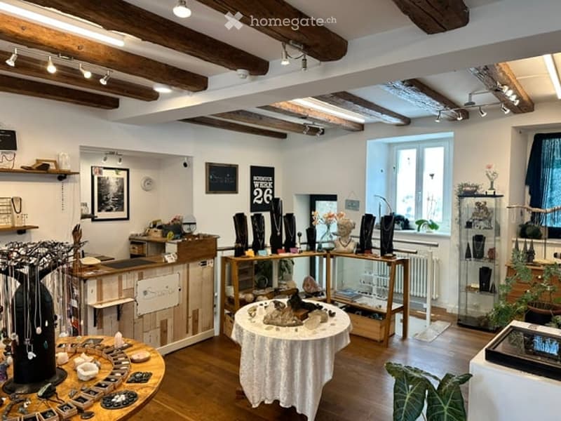 A well-lit boutique interior with exposed wooden beams, white walls, and wooden flooring. Shelves and display cases hold various decorative items, including vases, necklaces, and artworks. A central round table with a white tablecloth displays additional