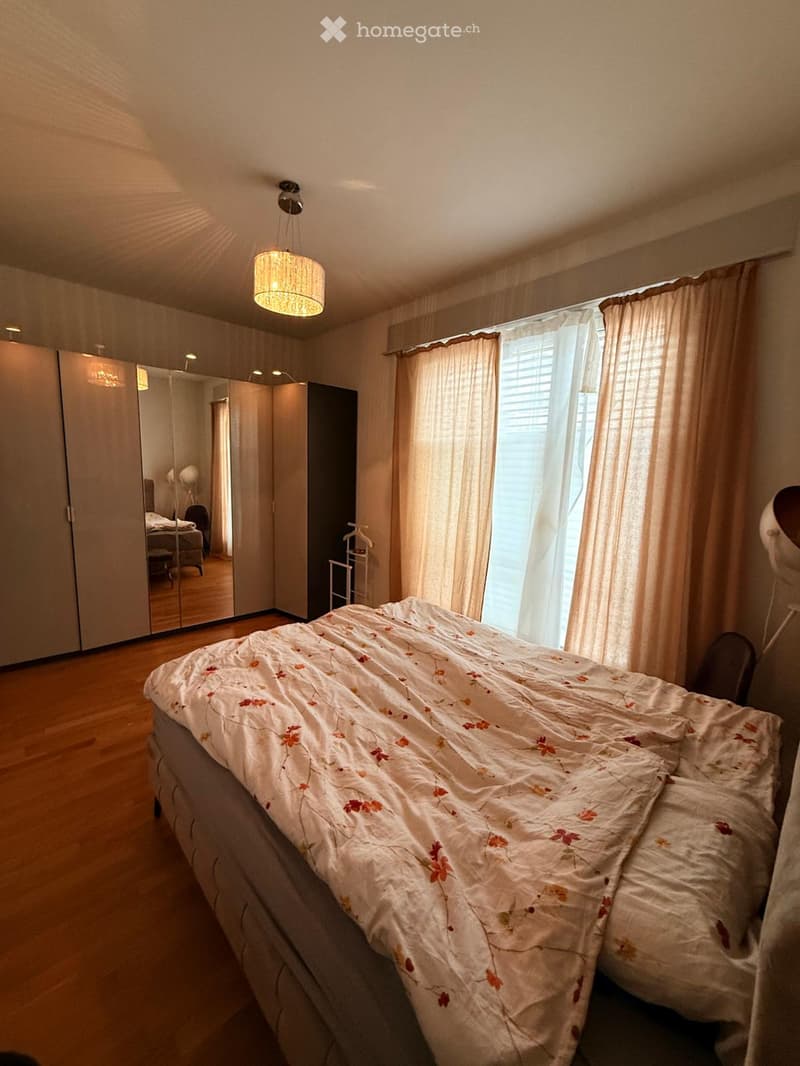 A well-lit bedroom with a wooden floor, a large bed with a floral duvet, a wardrobe with mirrored doors, and large windows with curtains.