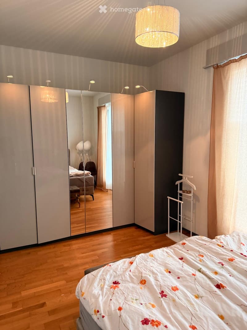 Modern bedroom with wooden flooring, large mirrored wardrobe, bed with floral duvet, and a hanging lamp.