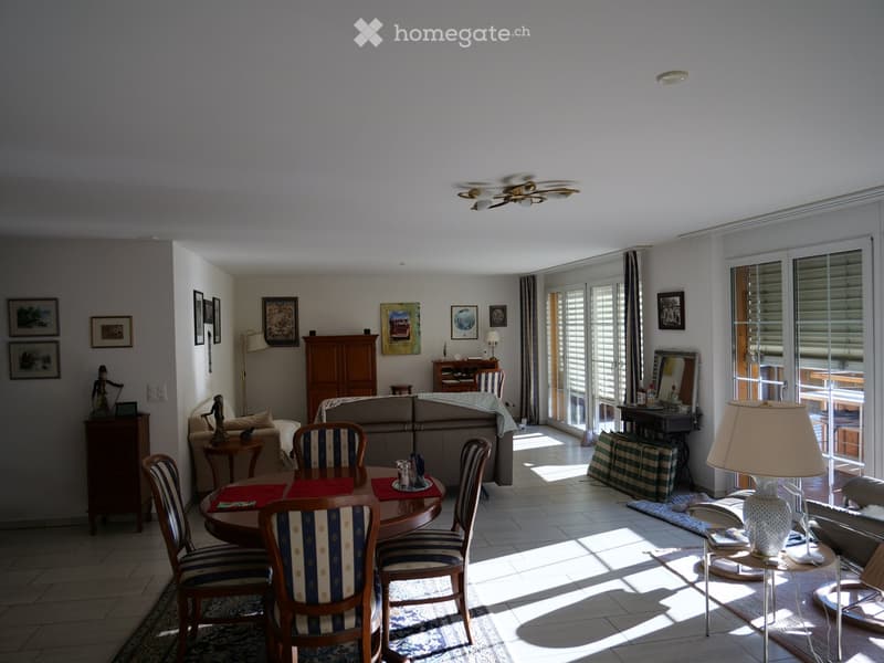 Bright living room with a sofa, dining table, chairs, lamps, and artwork on the walls. Large windows and sliding glass doors lead to a balcony area.