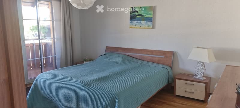 Cozy bedroom with a wooden bed, teal bedding, a nightstand with a lamp, and a painting on the wall. Large window with a view of the outdoor area.