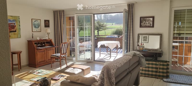 Bright living room with large sliding glass doors leading to a balcony, sofa, coffee table, desk, chair, mirror, and a small table with a white tablecloth on the balcony.