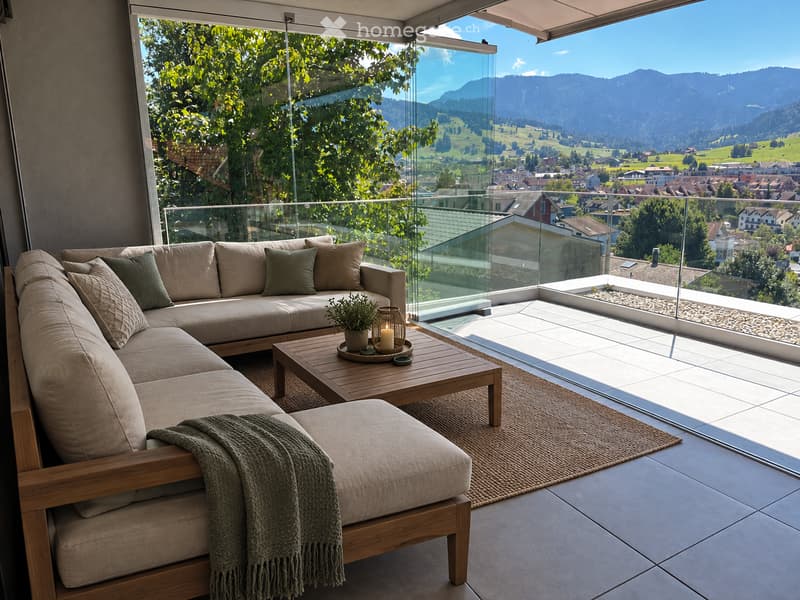 Modern balcony with panoramic mountain view, featuring a beige sectional sofa, wooden coffee table, ottoman, and decorative elements.