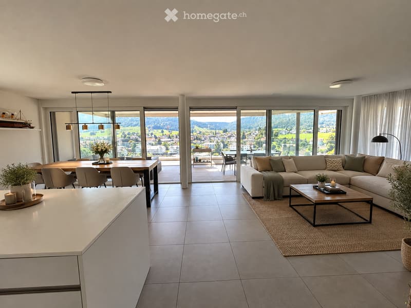 Modern open-plan living and dining area with large windows offering scenic views, featuring a sectional sofa, coffee table, dining table with chairs, and decorative elements like candles and plants.