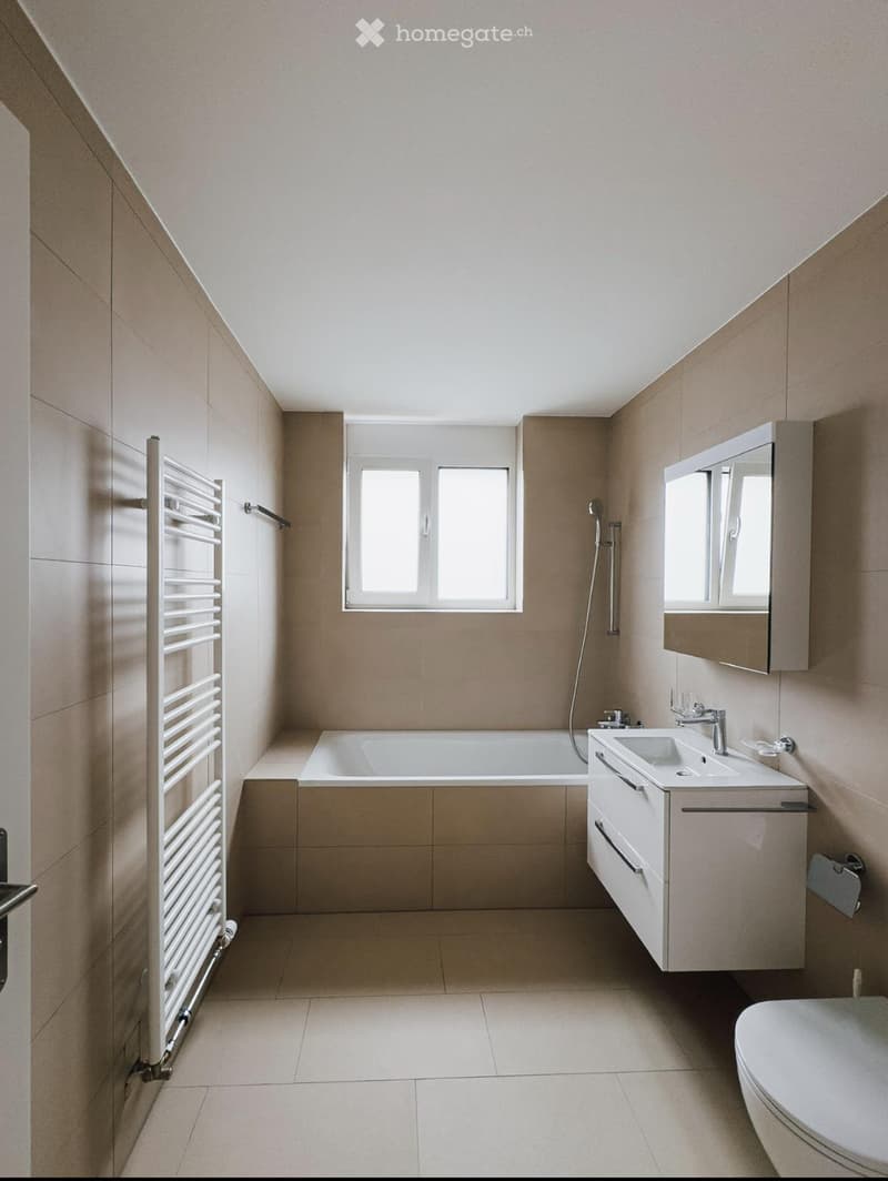 Modern bathroom with beige tiles, a bathtub, a sink with a cabinet, a toilet, a mirror, and a radiator.
